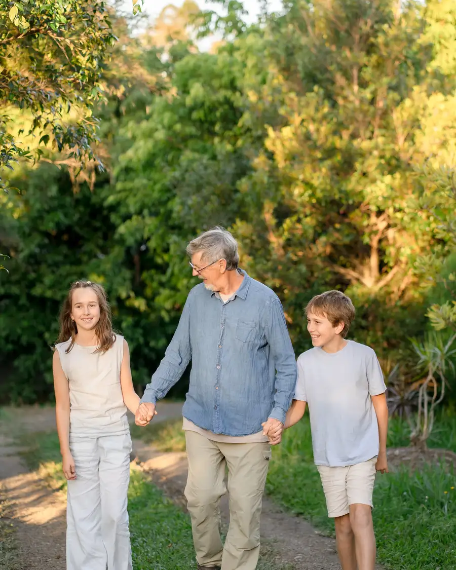 A grandfather walking hand in hand with two children along a sunlit garden path