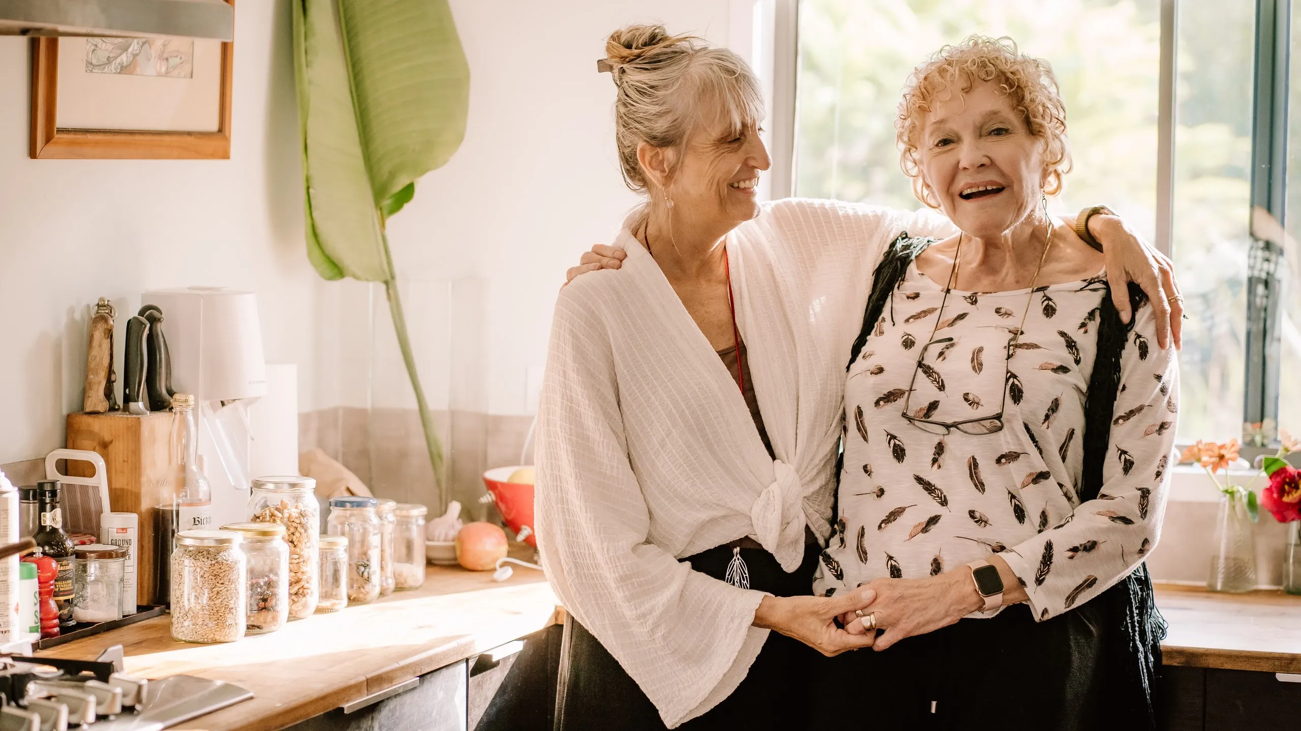 Two older women sharing a warm moment together in a bright, sunlit kitchen